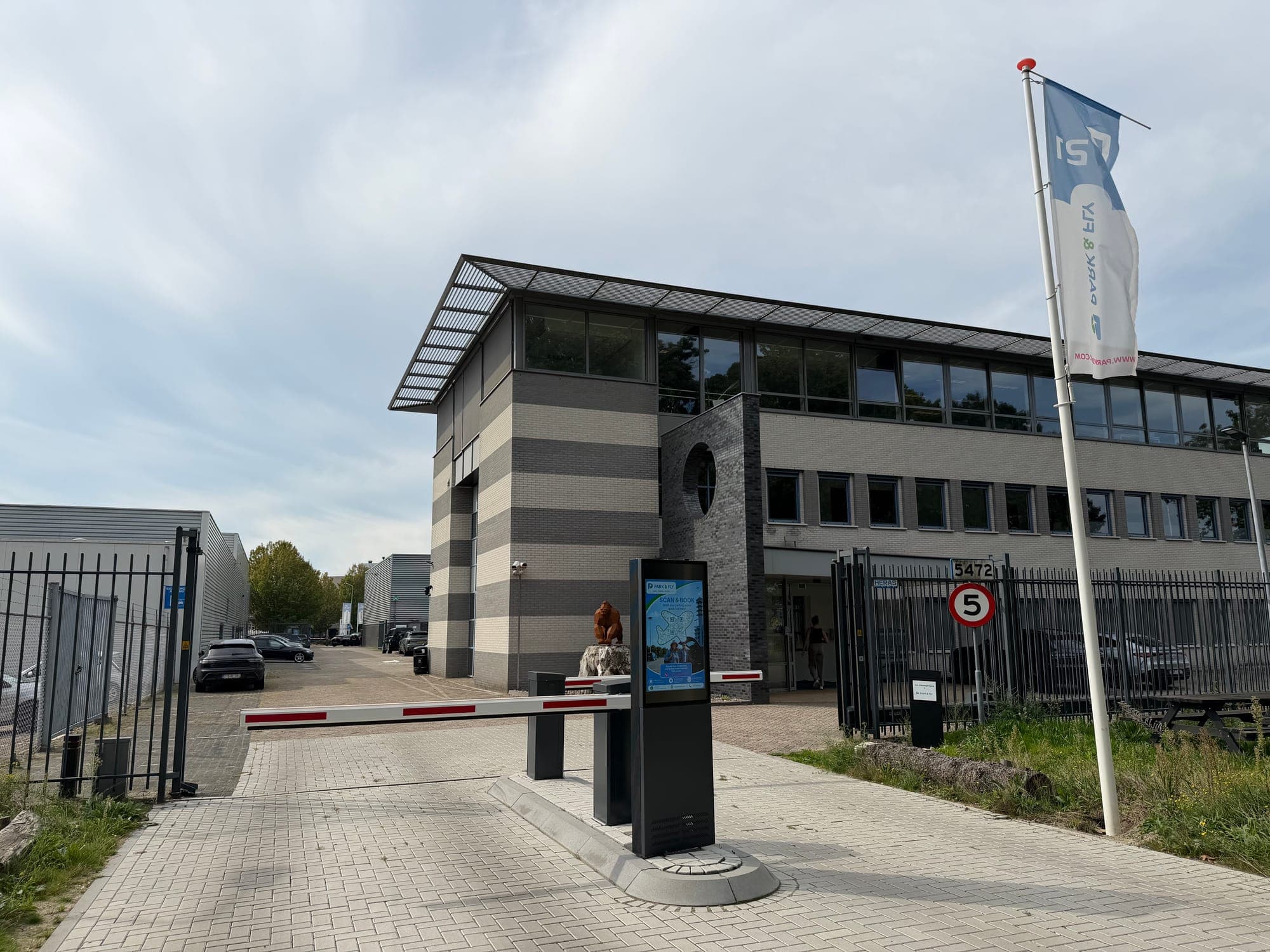 Die Zufahrt von Park en Fly P21 am Flughafen Eindhoven mit Schranke, Informationssäule und der P21-Fahne vor dem Büro.